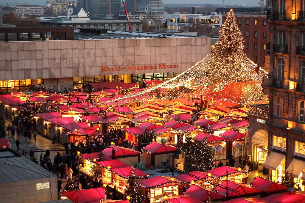 Rows of red-roofed stalls and a lit Christmas tree at the Christmas market beside Cologne Cathedral at dusk