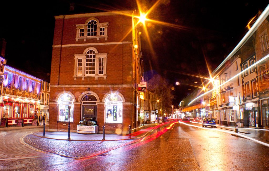 Market Harborough town centre on a winter evening with lit shop windows and the historic red-brick corner building on The Square