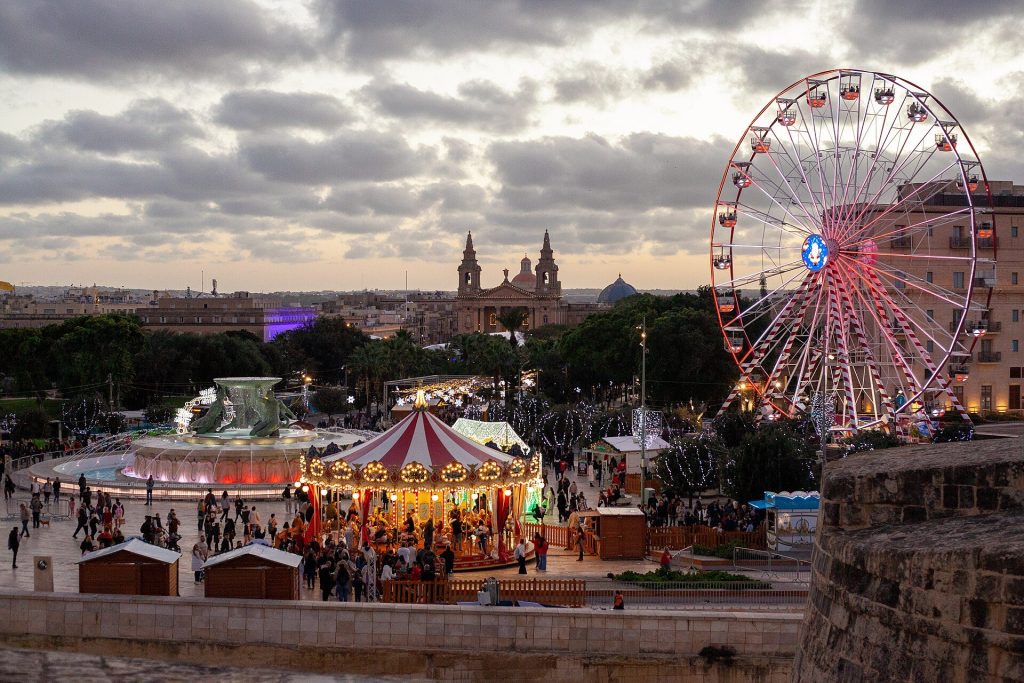 Christmas market chalets, a carousel and a Ferris wheel around the Triton Fountain outside City Gate in Valletta at dusk