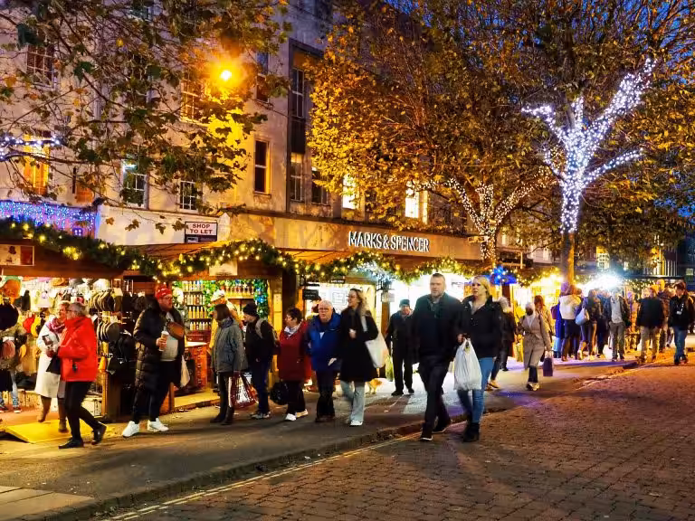 Shoppers walking past garland-lit chalets at York Christmas Market on Parliament Street at dusk